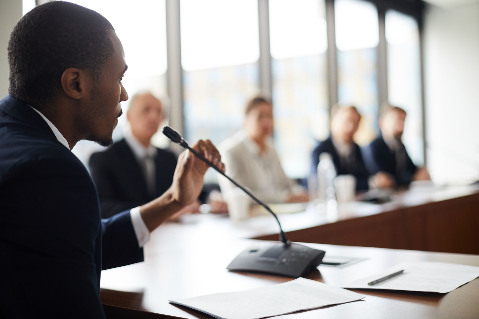 Serious confident black businessman sitting at conference table with papers and holding microphone while expressing his viewpoint at press meeting Serious confident black businessman sitting at conference table with papers and holding microphone while expressing his viewpoint at press meeting